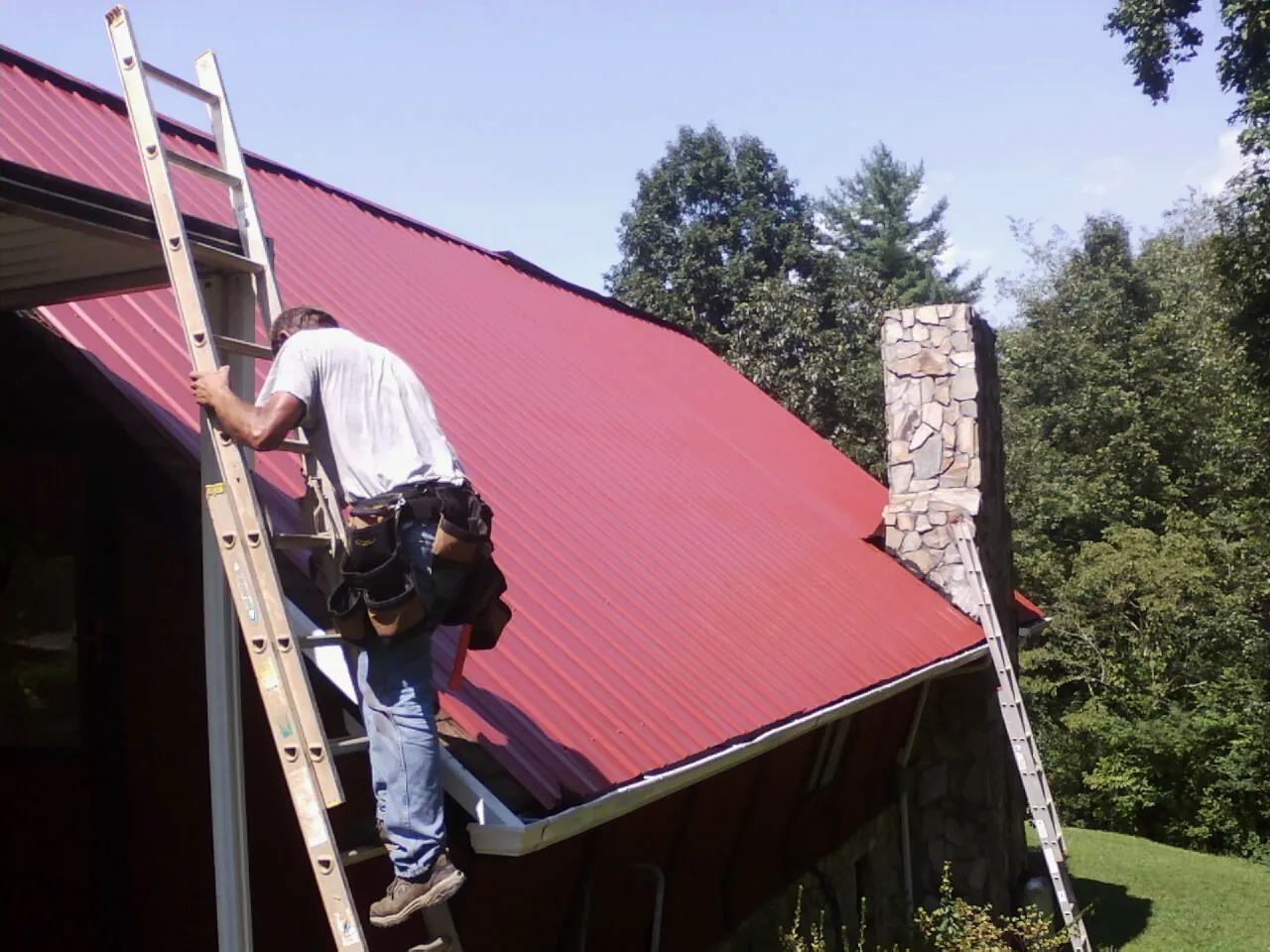 Roofer on a ladder beside a red metal roof and stone chimney — Soffit & Fascia Repair by Roofing Brothers in Irene